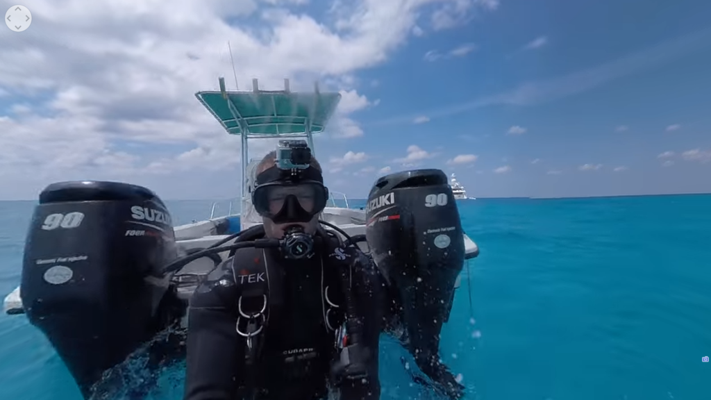 Audio Description: National Geographic logo. On tropical waters, you and another scuba diver stand at the edge of your boat and jump into the ocean. Onscreen text says: “Bimini, Bahamas.”