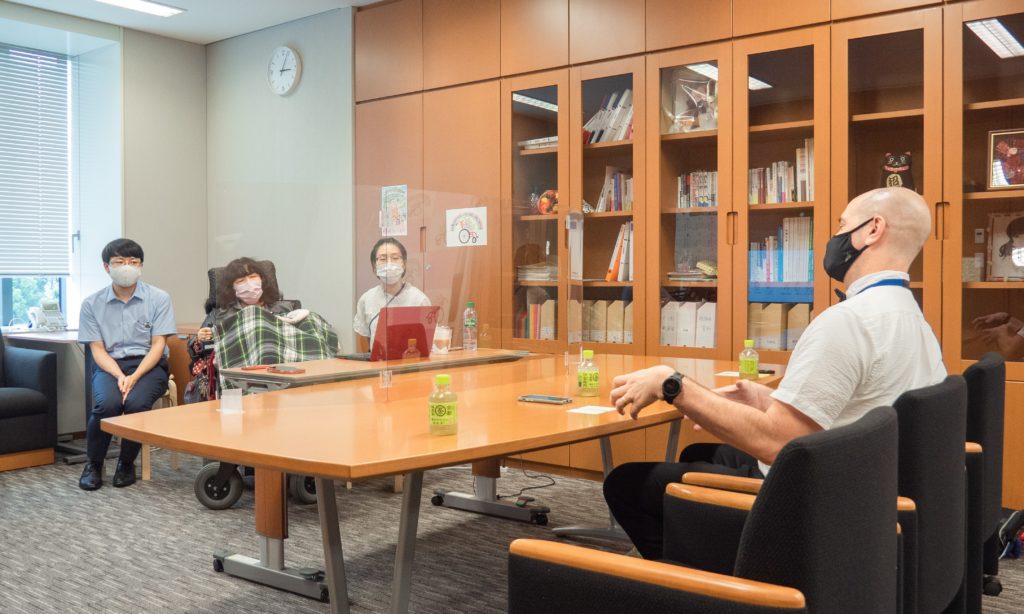 Thomas Logan sits across from Eiko Kimura who is in a wheelchair flanked by her aid and another person