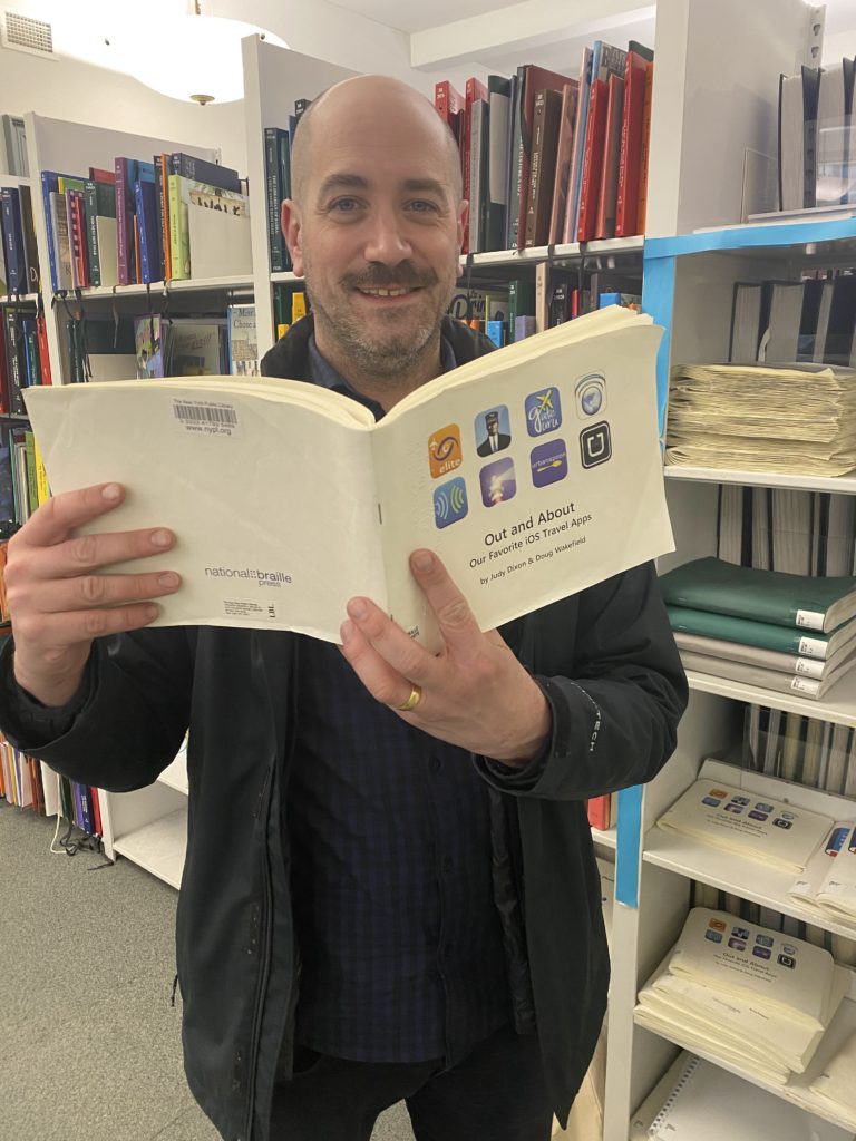 Thomas smiling while holding "Out and About: Our Favorite iOS Travel Apps" Braille book