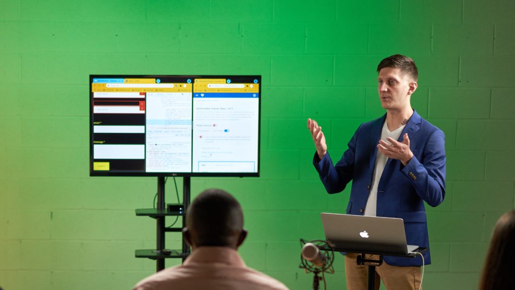 A medium-sized screen appears to the left of Cam who is talking while wearing a blue jacket over a white V-neck shirt and green wall behind