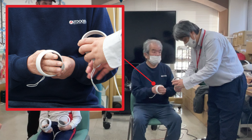 A senior user put their hand through the ring of the hand controller. Azalee staff shows him how to hold the controller.