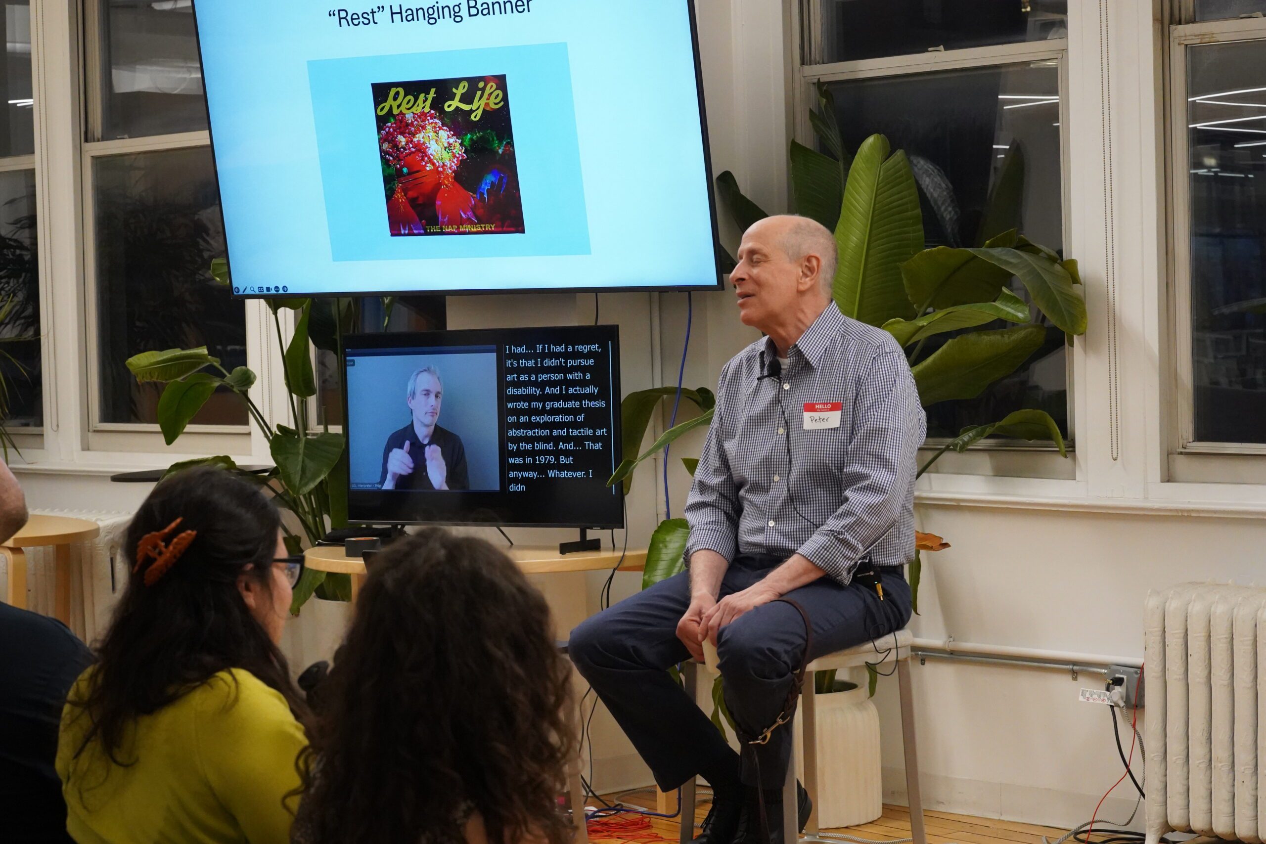 Peter Slatin sits on a stool next to a large screen with his slides and below the screen is a smaller screen displaying an ASL interpreter and captions.
